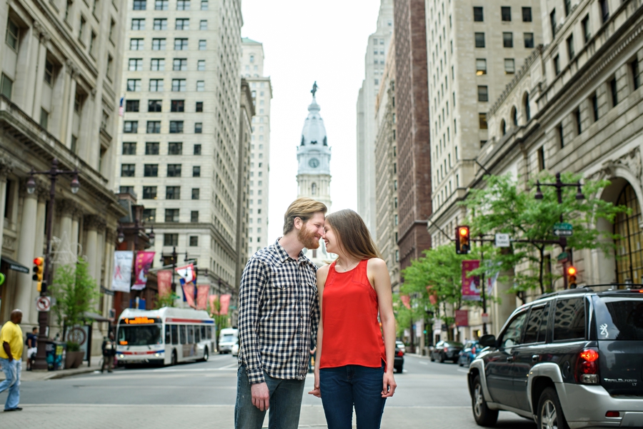 An engaged couple touch noses on broad street in philadelphia with city hall in the background.